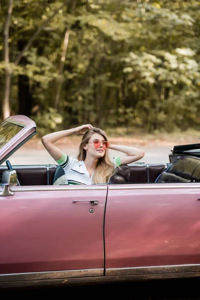 Femme souriante dans des lunettes de soleil toucher les cheveux et regarder loin tout en étant assis dans le cabriolet vintage — Photo de stock