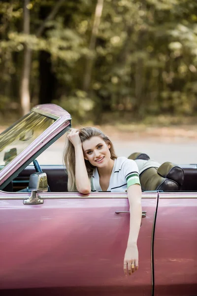 Femme joyeuse toucher les cheveux tout en étant assis dans une voiture convertible et en regardant la caméra — Photo de stock