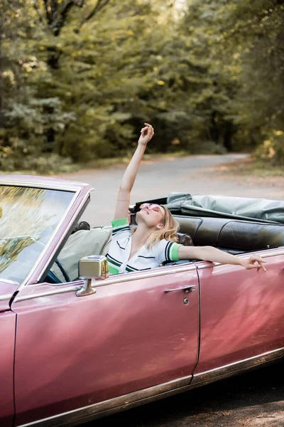 Joyful woman relaxing in cabriolet while sitting with raised head, closed eyes and hand in air — Stock Photo