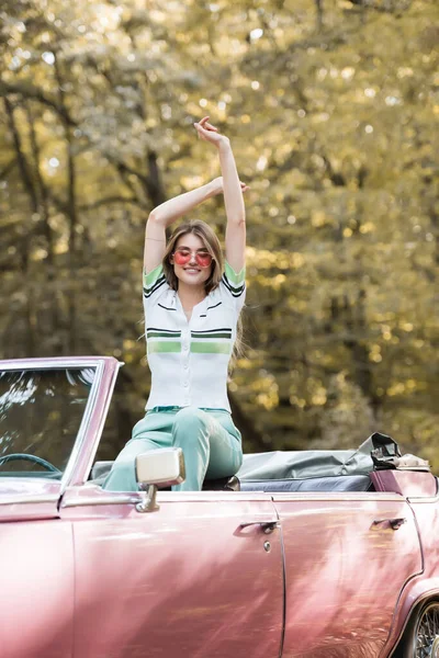 Femme joyeuse dans des lunettes de soleil assis dans une voiture convertible avec les mains levées — Photo de stock