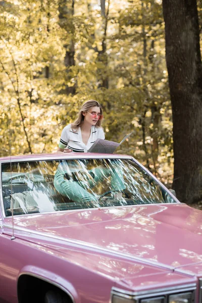 Junge Frau sitzt im Cabrio und schaut im Wald auf Landkarte — Stockfoto