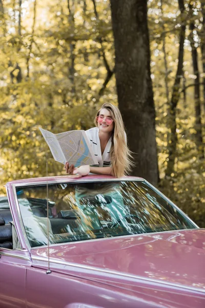 Femme souriante dans des lunettes de soleil regardant atlas de la route tout en étant assis dans le cabriolet dans la forêt — Photo de stock