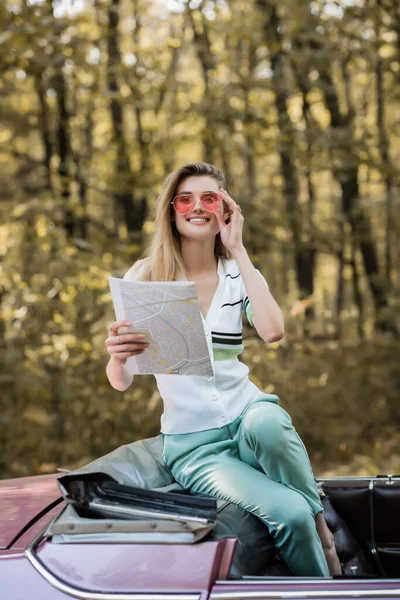Cheerful woman touching sunglasses and looking at camera while holding map in cabriolet — Stock Photo