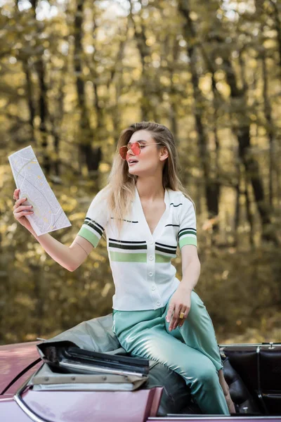 Stylish woman in sunglasses looking at road atlas while sitting in cabriolet in forest — Stock Photo