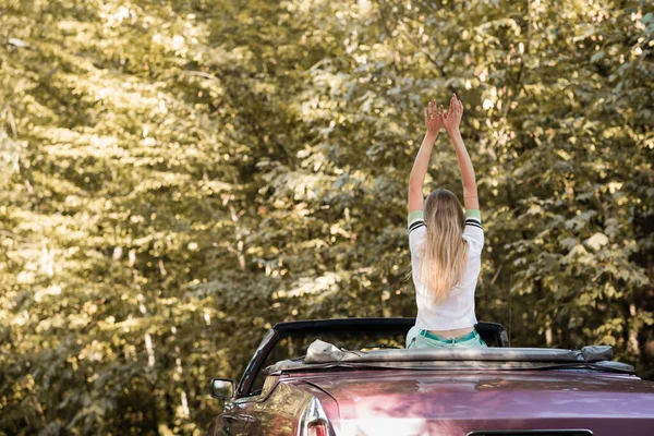 Vue arrière de la jeune femme assise dans le cabriolet avec les mains levées dans la forêt — Photo de stock