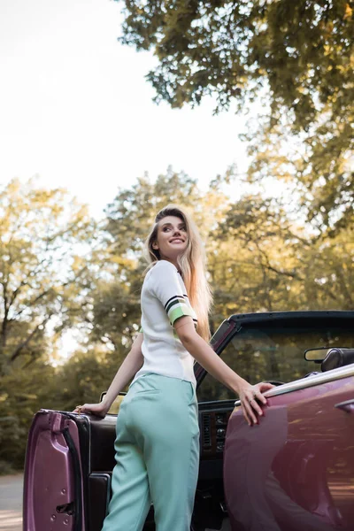 Joyeuse jeune femme regardant loin tout en ouvrant la porte du cabriolet vintage — Photo de stock