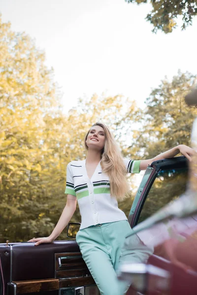 Low angle view of smiling woman looking away while standing near open door of cabriolet on blurred foreground — Stock Photo