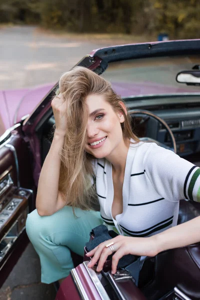 Gioiosa giovane donna toccare i capelli e guardando la fotocamera mentre seduto in auto convertibile — Foto stock
