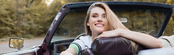 Mujer sonriente mirando hacia otro lado mientras está sentado en un coche descapotable, pancarta - foto de stock