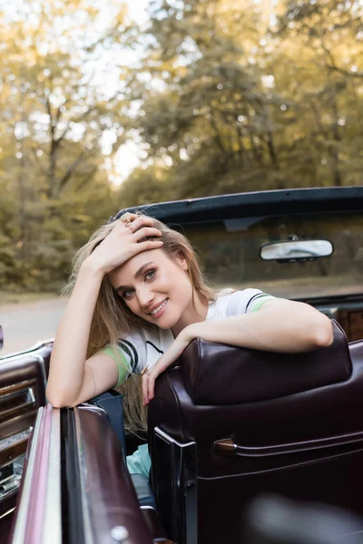 Donna allegra seduta in cabriolet vintage, toccando i capelli e guardando la fotocamera, in primo piano sfocato — Foto stock
