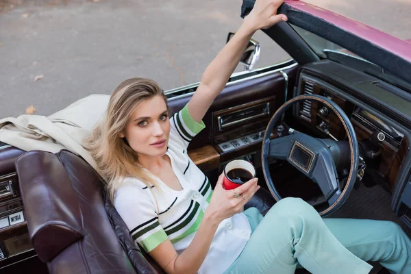 High angle view of young woman looking at camera while sitting in cabriolet and holding cup of coffee — Stock Photo