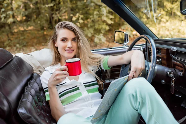 Jeune femme souriante regardant la caméra tout en étant assis dans une voiture convertible avec atlas de la route et tasse de café — Photo de stock