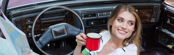 Femme gaie regardant loin tout en étant assis dans la voiture avec tasse de café et feuille de route, bannière — Photo de stock