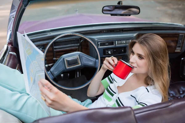 Young woman drinking coffee and looking at road atlas in cabriolet on blurred foreground — Stock Photo
