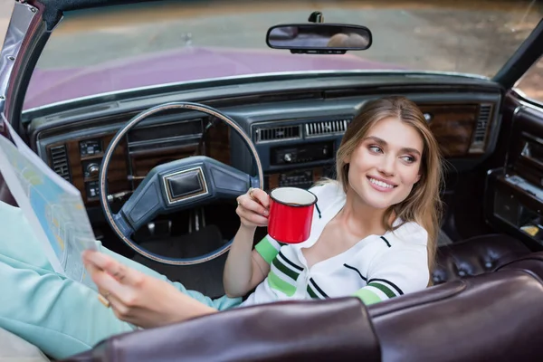 Mujer alegre mirando hacia otro lado mientras sostiene café y atlas carretera en cabriolet en primer plano borrosa - foto de stock