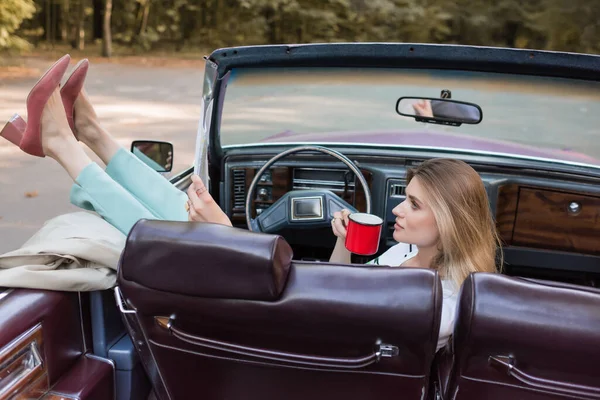 Frau legt Beine auf Tür, während sie Tasse Kaffee im Cabriolet hält — Stockfoto