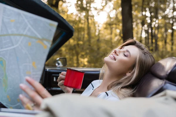Happy woman with closed eyes holding coffee and road atlas in cabrio on blurred foreground — Stock Photo