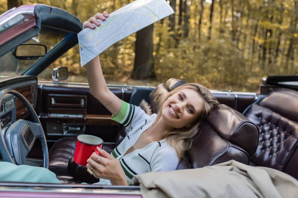 Aufgeregte Frau blickt in die Kamera, während sie Kaffee und Straßenatlas im Cabrio in der Hand hält — Stockfoto