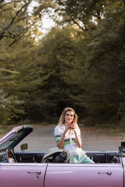 Young woman looking at camera while holding cup of coffee in cabriolet on road near forest — Stock Photo