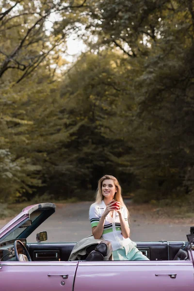 Lächelnde Frau mit Tasse Kaffee und Blick weg vom Oldtimer-Cabriolet in der Nähe des Waldes — Stockfoto