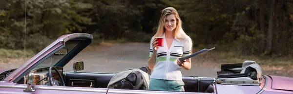 Donna sorridente che guarda la macchina fotografica mentre tiene atlante stradale e tazza di caffè in cabriolet, banner — Foto stock