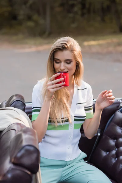 Stylische Frau trinkt Kaffee, während sie im Vintage-Cabriolet auf verschwommenem Vordergrund sitzt — Stockfoto