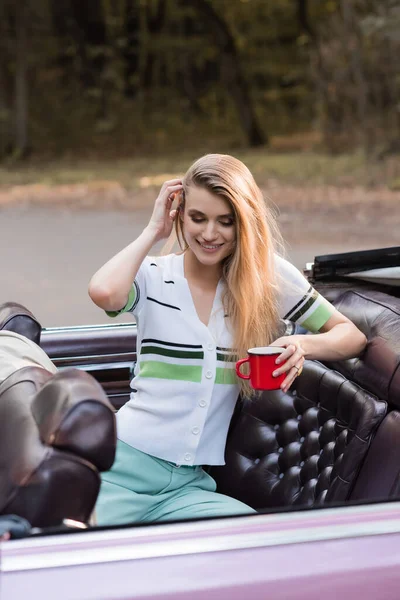 Улыбающаяся женщина касается волос, держа чашку кофе в винтажном кабриолете — стоковое фото