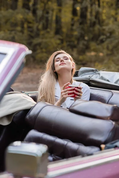 Mujer joven relajante mientras sostiene la taza de café con los ojos cerrados en cabriolet vintage en primer plano borroso - foto de stock
