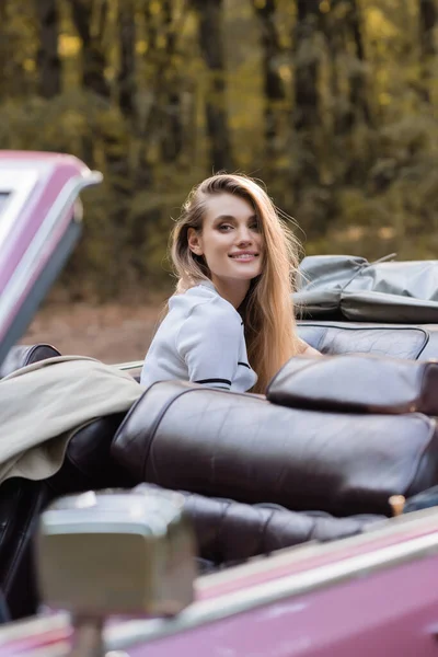 Молодая улыбающаяся женщина смотрит в камеру, сидя в кабриолете, размытый передний план — стоковое фото