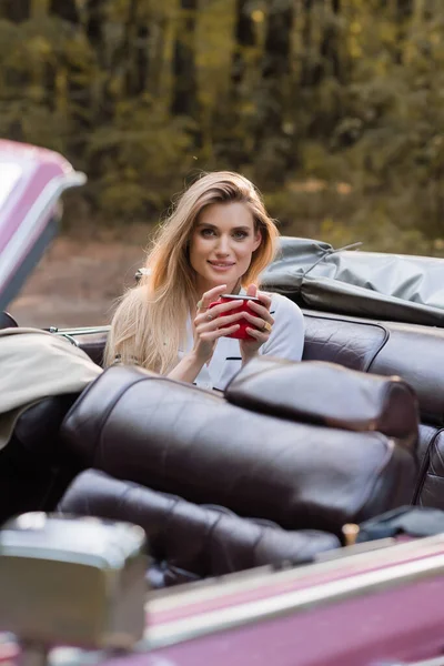 Young smiling woman looking at camera while sitting in cabriolet with cup of coffee on blurred foreground — Stock Photo