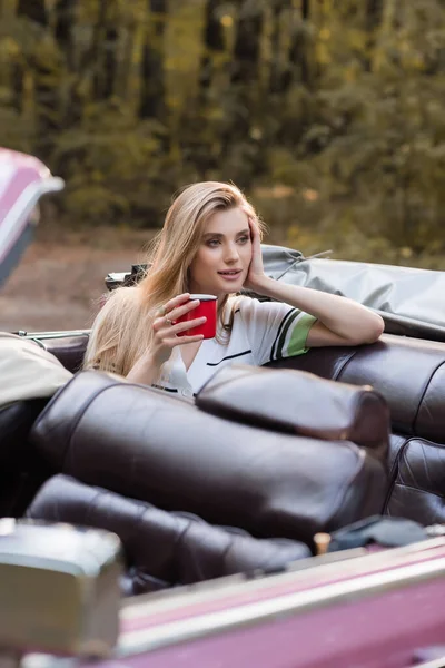 Mulher sorrindo segurando a mão perto do rosto e olhando para longe enquanto sentado em cabriolet com xícara de café em primeiro plano borrado — Fotografia de Stock