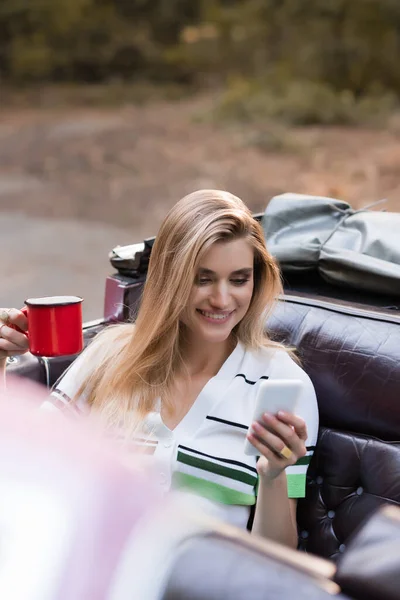 Donna allegra che tiene in mano una tazza di caffè e chatta sul telefono cellulare mentre siede in cabriolet su primo piano sfocato — Foto stock