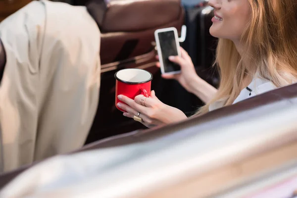 Vista parziale di giovane donna che tiene smartphone con schermo bianco e tazza di caffè in primo piano sfocato — Foto stock