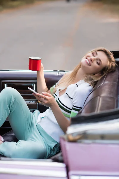 Happy woman holding book and smartphone while sitting in cabriolet with closed eyes on blurred foreground — Stock Photo