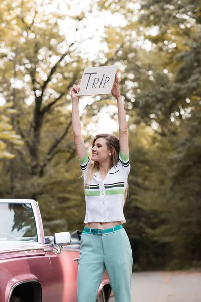 Young woman holding placard with trip lettering in raised hands while standing near vintage convertible car — Stock Photo