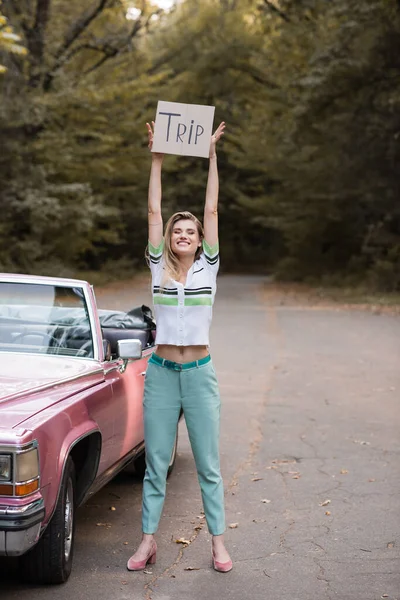 Junge lächelnde Frau blickt in die Kamera, während sie ein Plakat mit Trip-Schriftzug in der Nähe eines Oldtimer-Cabrios hält — Stockfoto