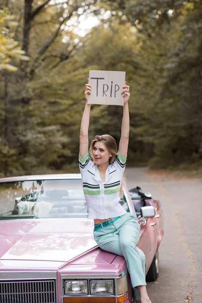 Smiling woman holding placard with trip lettering while sitting on cabriolet hood and looking away — Stock Photo