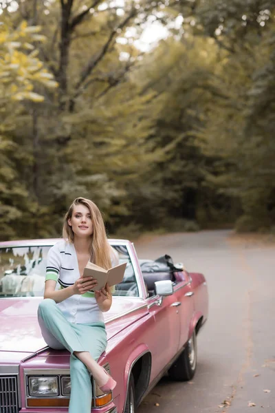 Femme souriante regardant la caméra tout en étant assis sur le capot du cabriolet et tenant livre — Photo de stock