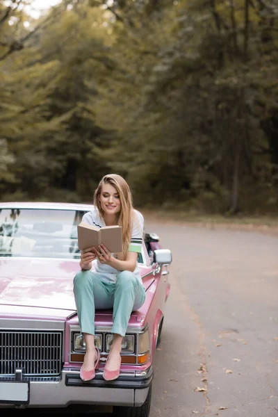 Mujer sonriente sentada en la capucha de cabriolet y libro de lectura - foto de stock