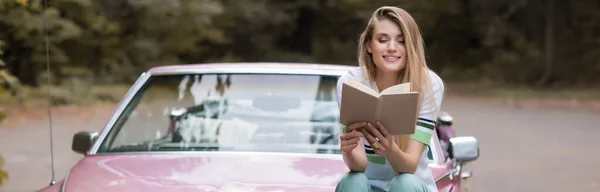 Весела молода жінка читає книгу, сидячи на капоті вінтажного кабріолета на розмитому фоні, банер — стокове фото