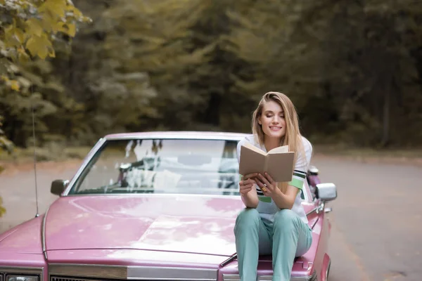 Heureuse jeune femme lecture livre tout en étant assis sur le capot de la voiture convertible sur fond flou — Photo de stock