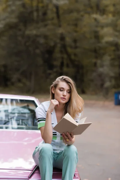 Nachdenkliche Frau sitzt auf der Motorhaube eines Cabrios und liest Buch auf verschwommenem Hintergrund, Banner — Stockfoto