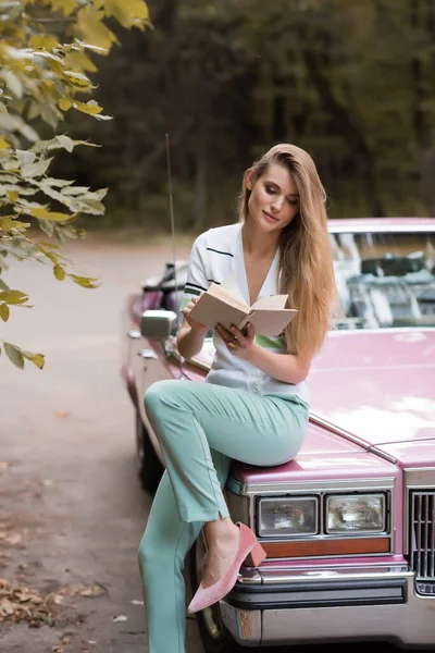 Stilvolle Frau liest Buch, während sie auf der Motorhaube eines Oldtimer-Cabrios sitzt — Stockfoto