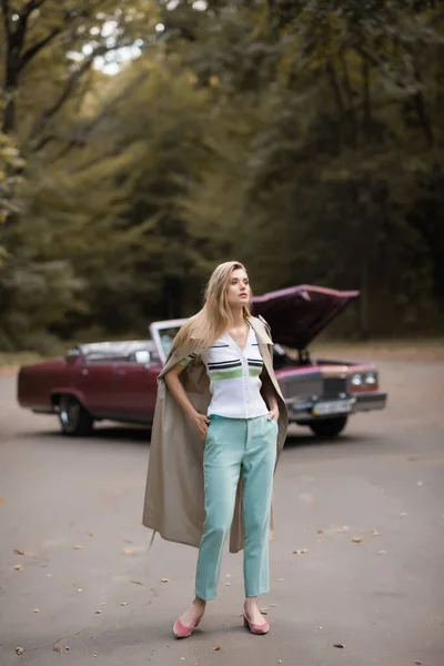 Femme élégante avec les mains dans les poches debout sur la route près cassé cabriolet sur fond flou — Photo de stock