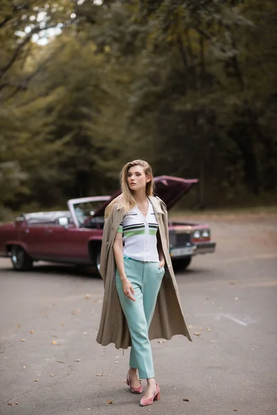 Mujer joven y elegante caminando con la mano en el bolsillo cerca de cabriolet roto con capucha abierta sobre fondo borroso - foto de stock