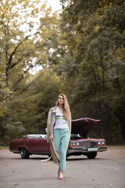 Mulher elegante na capa andando na estrada perto de carro vintage quebrado no fundo embaçado — Fotografia de Stock
