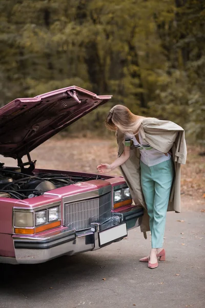 Mujer joven mirando bajo capucha abierta de coche vintage roto en la carretera - foto de stock