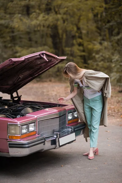 Jeune femme regardant sous le capot ouvert de voiture vintage cassée sur la route — Photo de stock