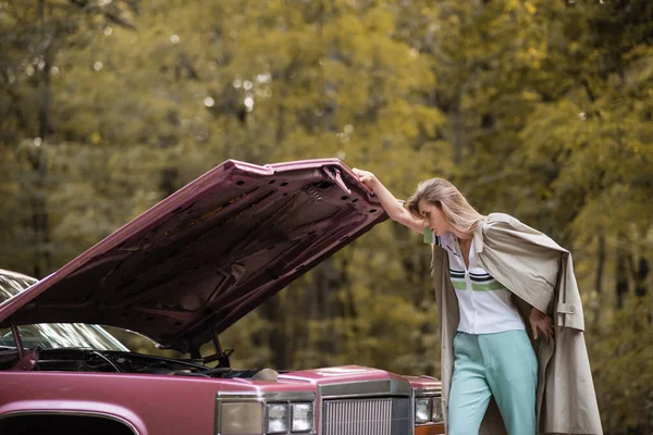Young woman in cape looking under open hood of broken vintage car — Stock Photo
