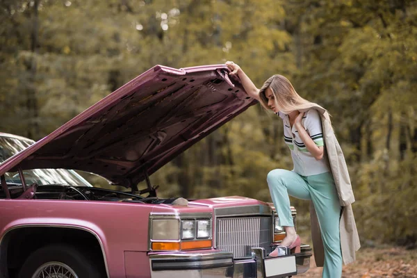 Young stylish woman looking under open hood of broken retro car — Stock Photo